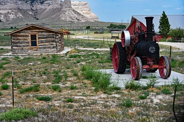 Historical Heritage at the Edge of the Prairie – Steam tractor and log cabin in front of the bright cliffs of Scotts Bluff. nz7 8999